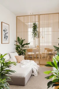 Tropical Modern Apartments A cozy, sunlit living space with a daybed, several potted plants, and a slatted room divider separating the area from a dining table and chairs.