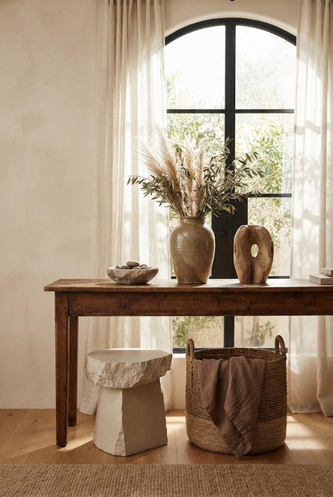 A wooden console table with a ceramic vase of dried grasses, a stone bowl and a carved wooden sculpture in front of an arched window, with a stone stool and woven basket below.